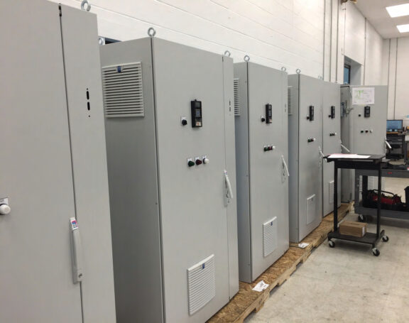 Row of tall gray electrical control cabinets with panels, vents, and control buttons in an industrial room on a wooden platform.