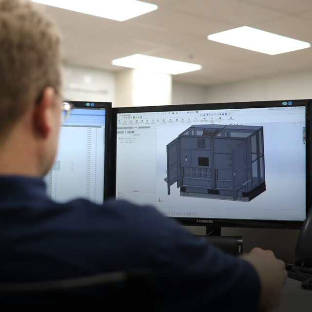 Worker at a desk viewing a 3D CAD model of an industrial enclosure on a monitor