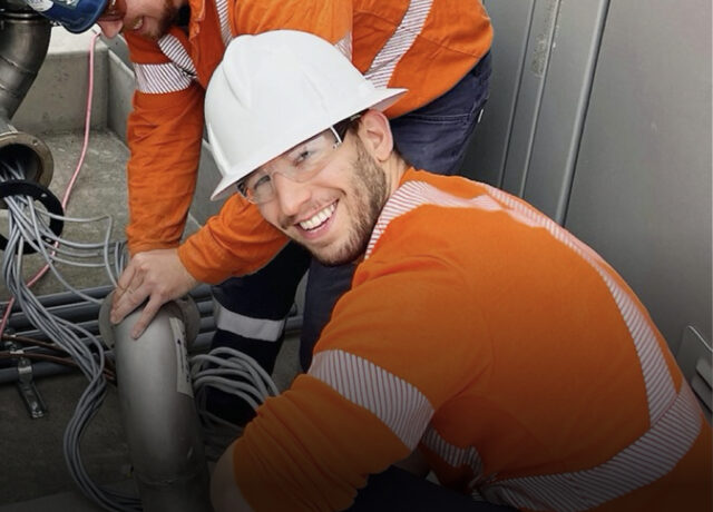 Two construction workers in orange safety jackets and white hard hats manage cables on a concrete floor, smiling at the camera.