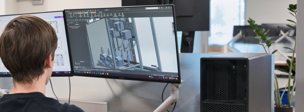 Person seated at a desk, viewing a 3D CAD model of machinery on a large monitor with a desktop PC nearby and a plant in the background